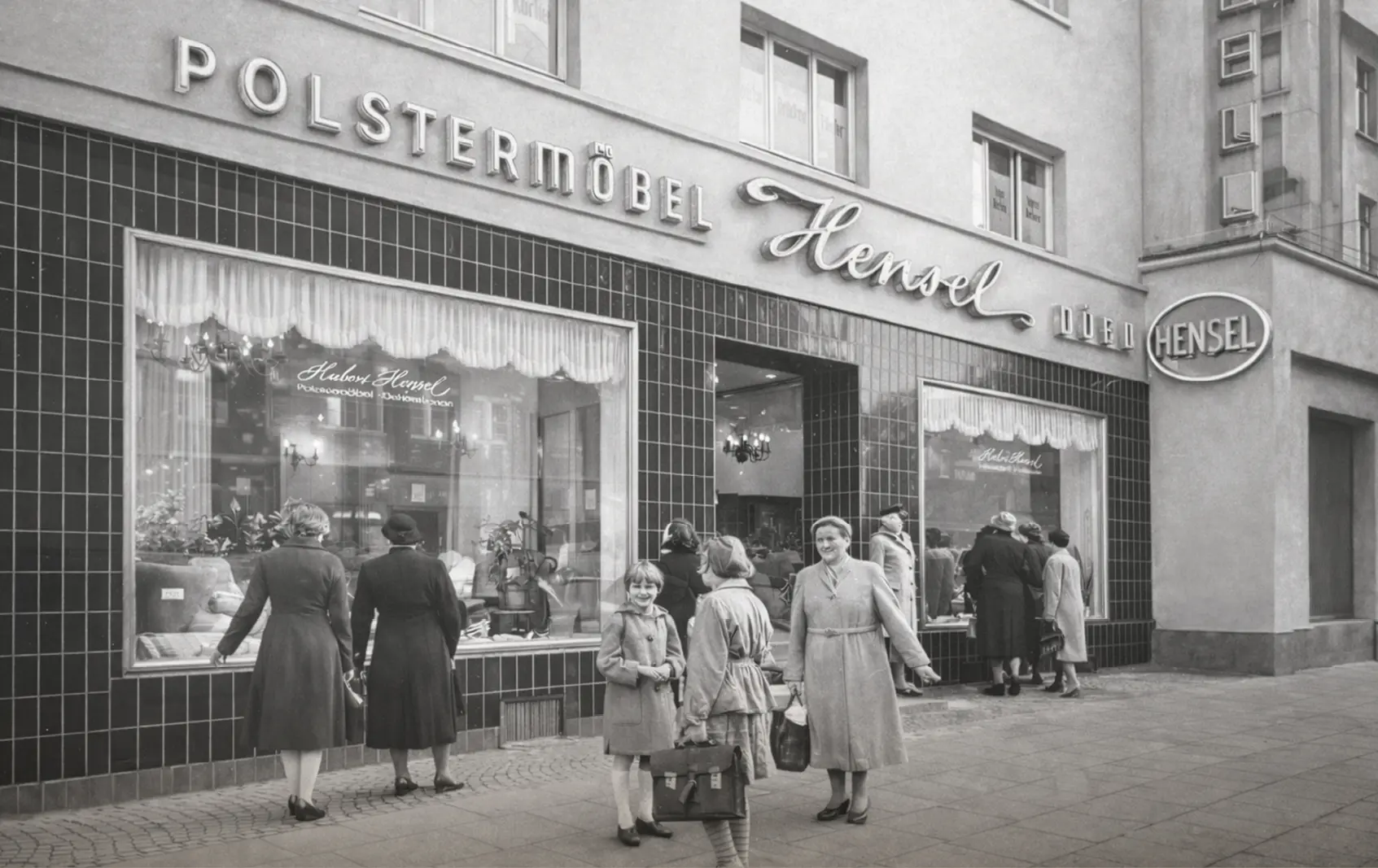 Schwarzweißaufnahme eines Möbelgeschäfts mit Schriftzug Polstermöbel Hensel, Passanten stehen vor großen Schaufenstern